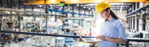 Frau mit Helm in Fabrikhalle schaut auf Laptop