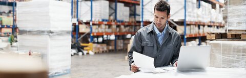 Mann in Fabrikhalle liest sich Unterlagen durch