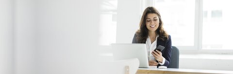 Frau mit Handy in der Hand vor dem Laptop