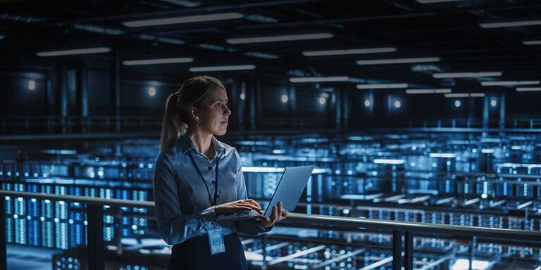Eine Frau mit Laptop kontrolliert etwas, im Hintergrund das Rechenzentrum in dunkelblauem Licht