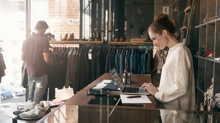 Eine Ladenbesitzerin steht an einem MacBook in einem kleinen Bekleidungsgeschäft, im Hintergrund ein Kunde
