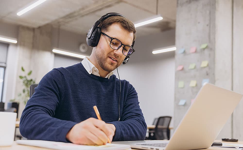 Ein Mann im blauen Pullover mit Headset hört bei einem Meeting zu und macht sich Notizen.