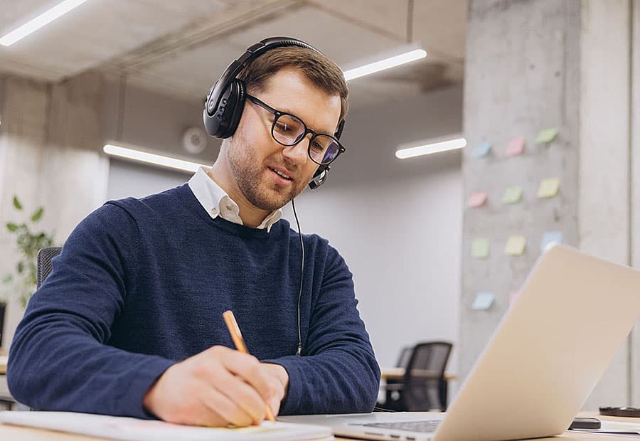 Ein Mann im blauen Pullover mit Headset hört bei einem Meeting zu und macht sich Notizen.