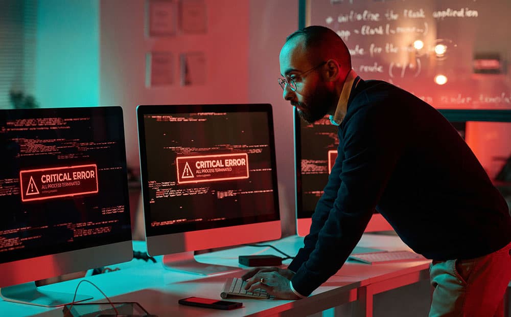 Ein Mitarbeiter vor zwei rot erleuchteten Bildschirmen, auf denen der Hinweis "Critical Error" zu lesen ist
