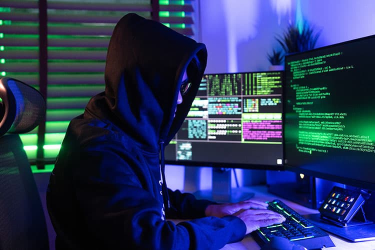 Ein Hacker mit Hoodie vor Bildschirmen mit Code in grüner Schrift