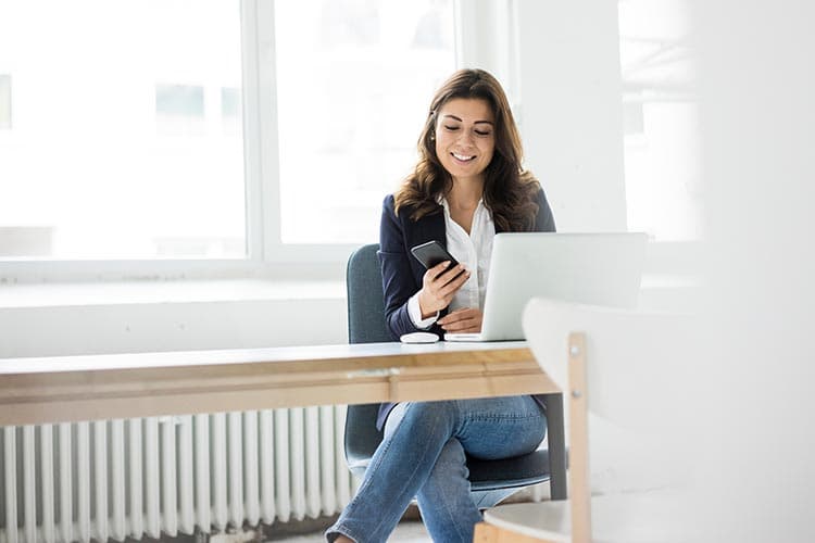 Eine Frau sitzt mit Handy vor Ihrem Arbeitsrechner und lächelt.