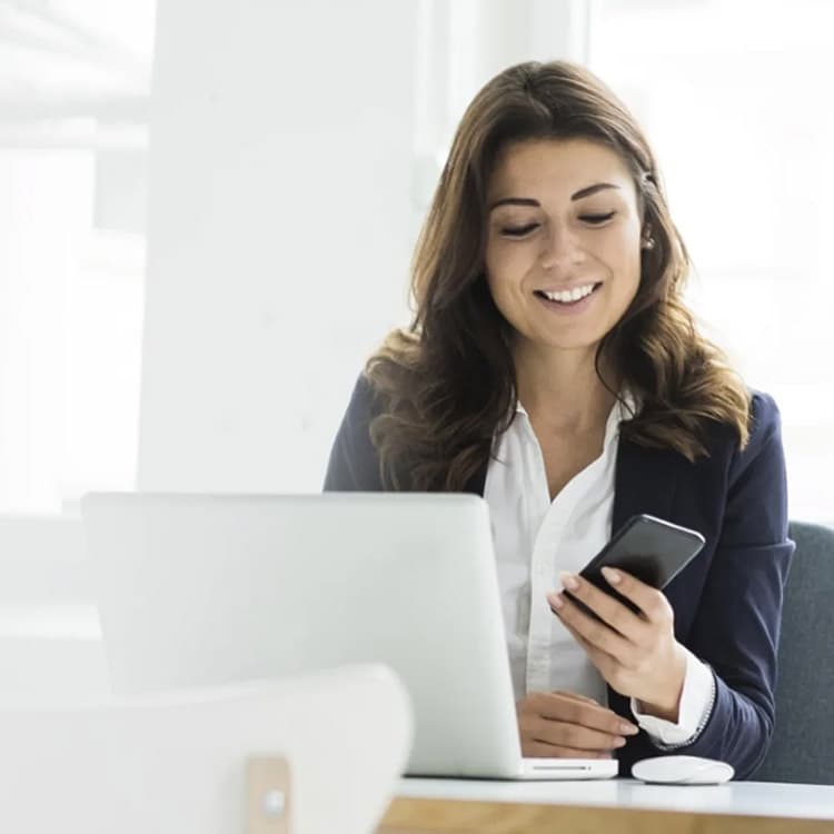 Eine Frau sitzt mit Handy vor Ihrem Arbeitsrechner und lächelt.