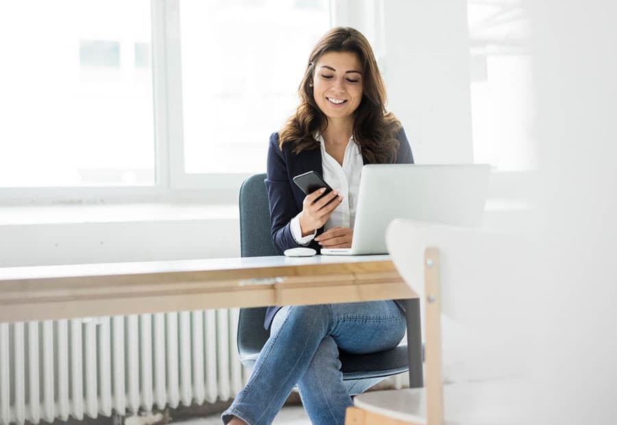 Eine Frau sitzt mit Handy vor Ihrem Arbeitsrechner und lächelt.