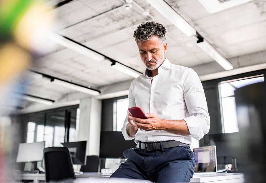 Ein Mann im weißen Hemd sitzt an einen Schreibtisch gelehnt und guckt etwas auf seinem Handy an.