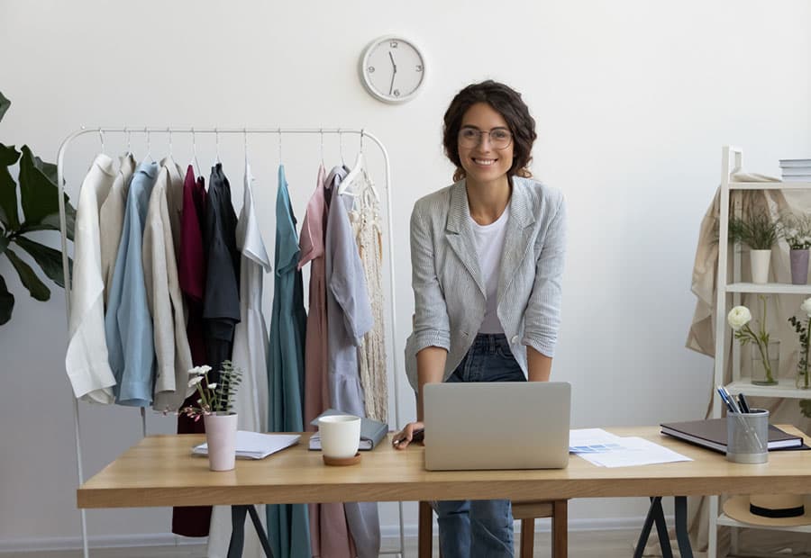 Eine junge Unternehmerin mit Arbeitstisch und Computer sowie Kleiderständer im Hintergrund 
