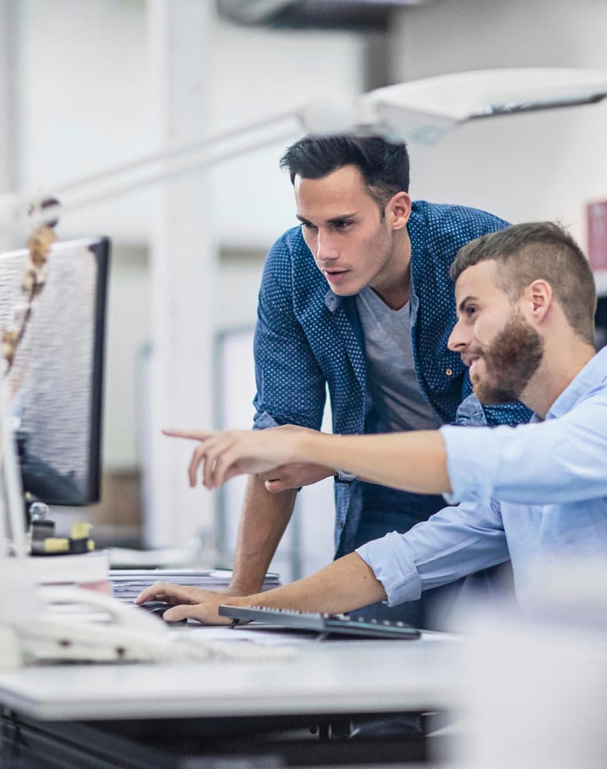 Zwei Männer besprechen im Büro etwas an einem Schreibtisch, der vordere zeigt auf einen Bildschirm.