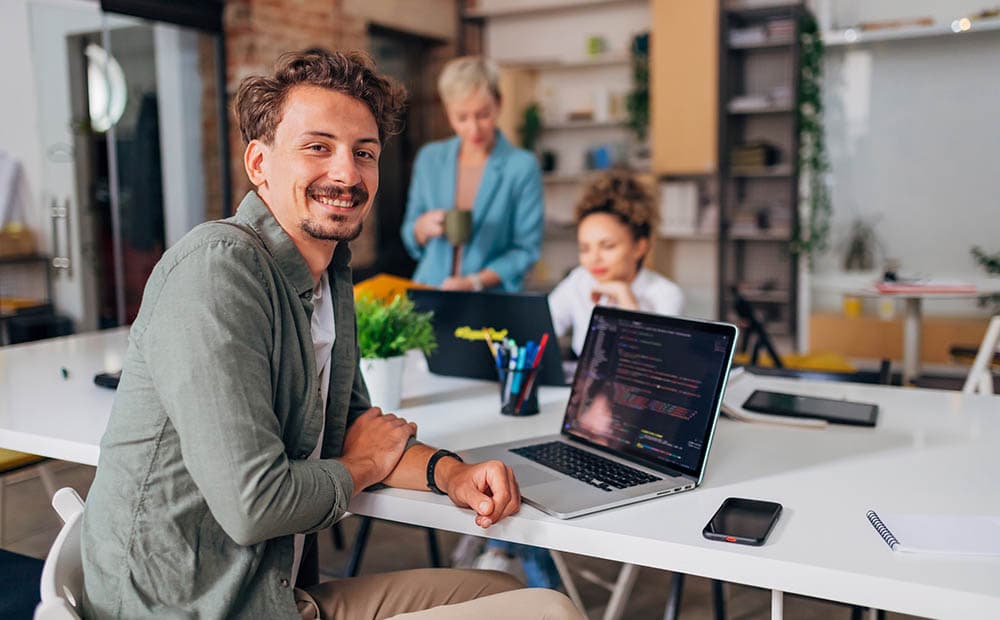 Ein Mann an einem Schreibtisch, der lächelt, dahinter zwei Kolleginnen