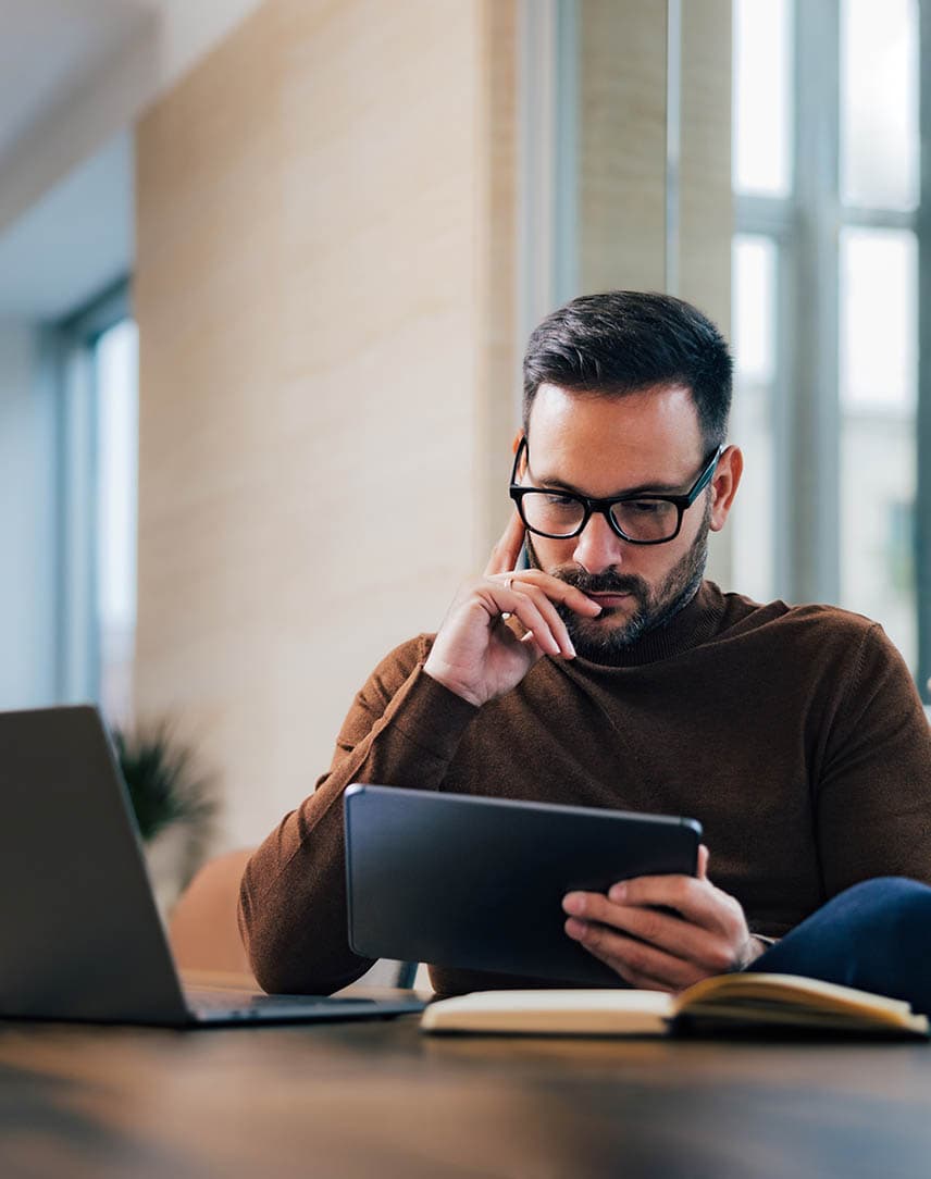 Ein Mann, der an seinem Schreibtisch mit Laptop nachdenkend auf sein Tablet guckt