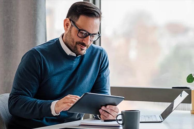 Ein Mann mit blauem Pullover an einem Büroschreibtisch, der auf ein Tablet guckt