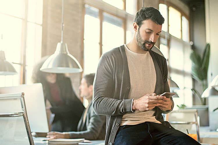 Mann sitzt auf Schreibtisch und schaut auf sein Smartphone