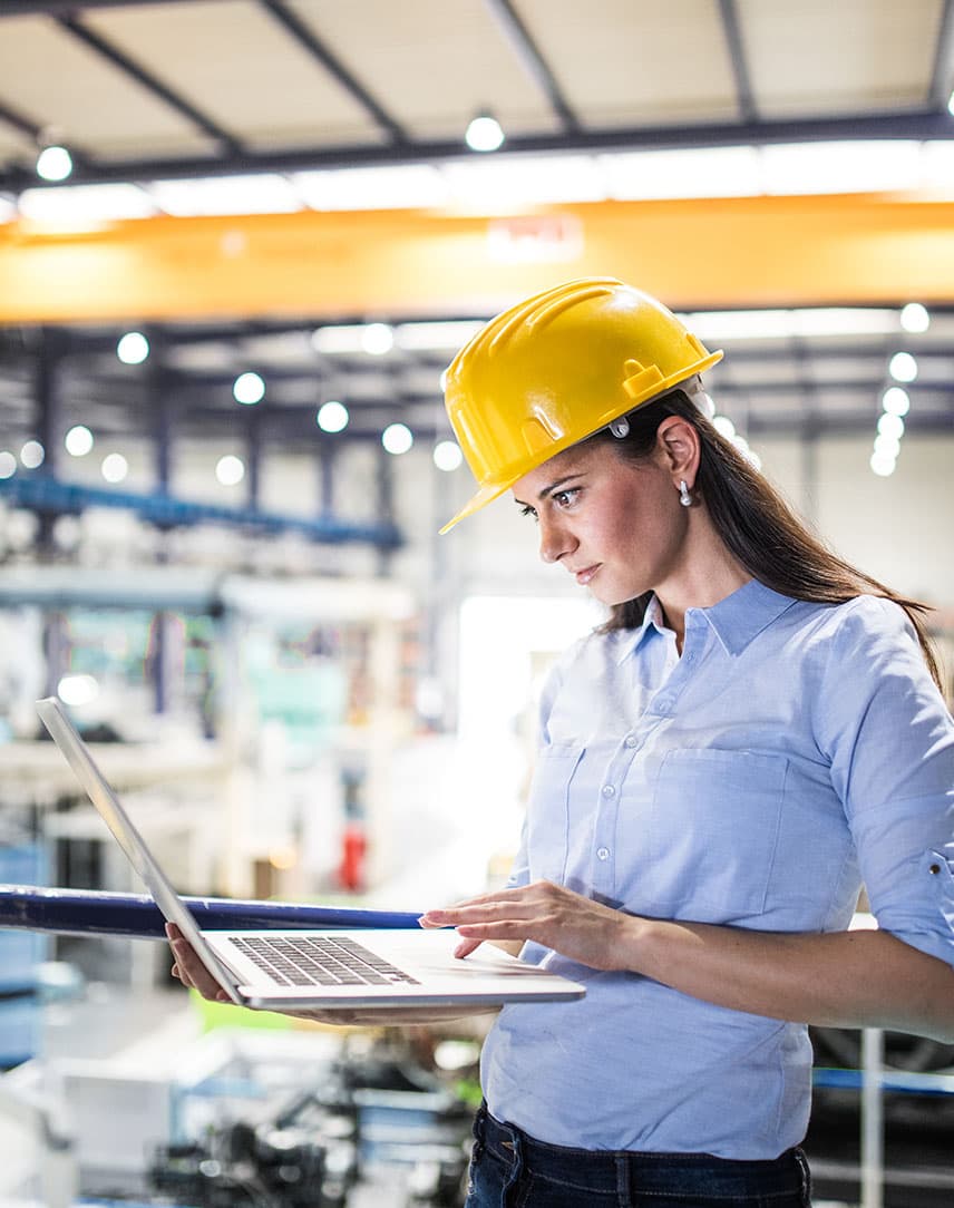 Frau mit Helm in Fabrikhalle schaut auf ihren Laptop.