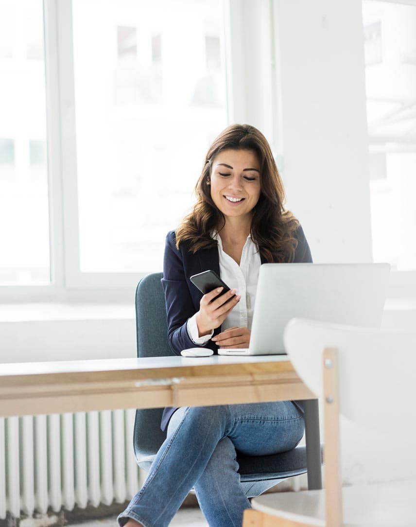 Eine Frau sitzt mit Handy vor Ihrem Arbeitsrechner und lächelt.