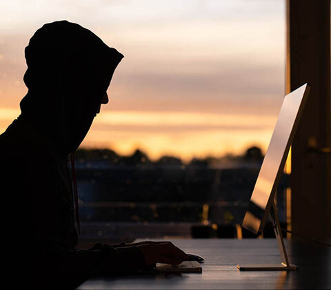 Stilisierter Hacker mit Kapuze über dem Kopf sitzt vor einem Computer