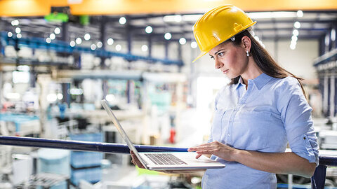 Frau mit Helm in Fabrikhalle schaut auf Laptop