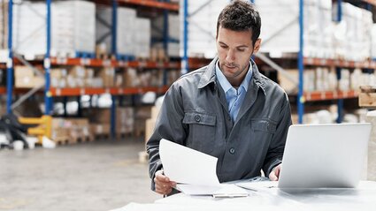 Mann in Fabrikhalle liest sich Unterlagen durch