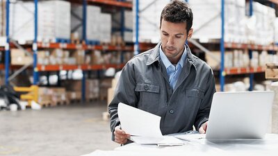Mann in Fabrikhalle liest sich Unterlagen durch