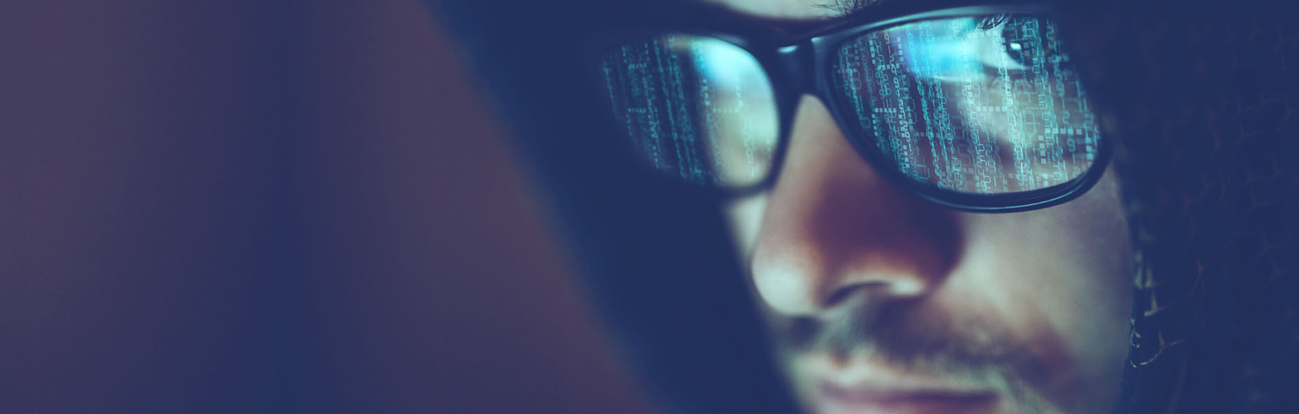 Close-up von einem Mann mit dunklem Hoodie und Brille, in der sich Computercode spiegelt.
