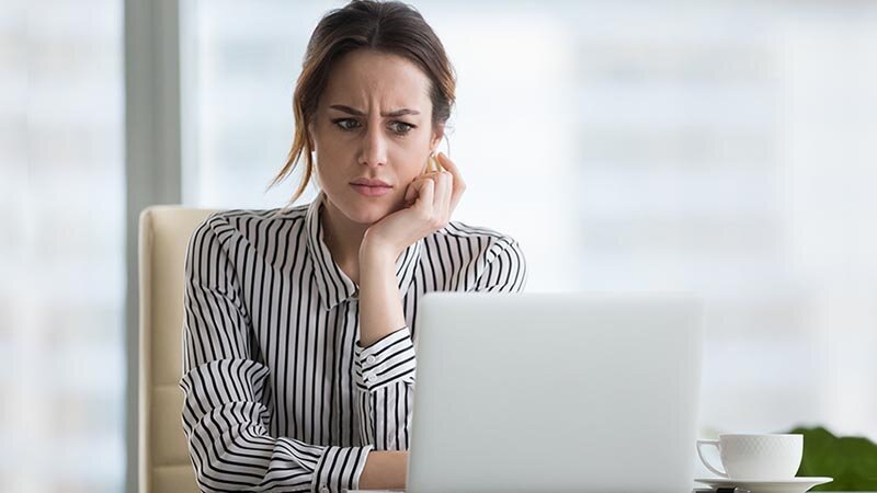 Eine dunkelhaarige Frau mit gestreifter Bluse sitzt am Schreibtisch und guckt zweifelnd auf ihren Computerbildschirm.