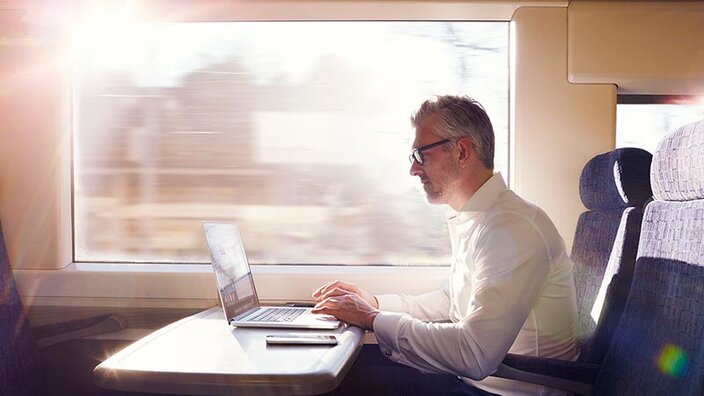 Ein Mann im weißen Hemd im Zug mit Laptop und Smartphone auf dem Tisch