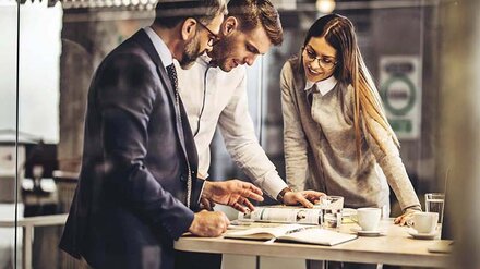 Zeitschriftenabo Drei KollegInnen, die um einen hohen Bürotisch stehen und auf eine Zeitschrift gucken